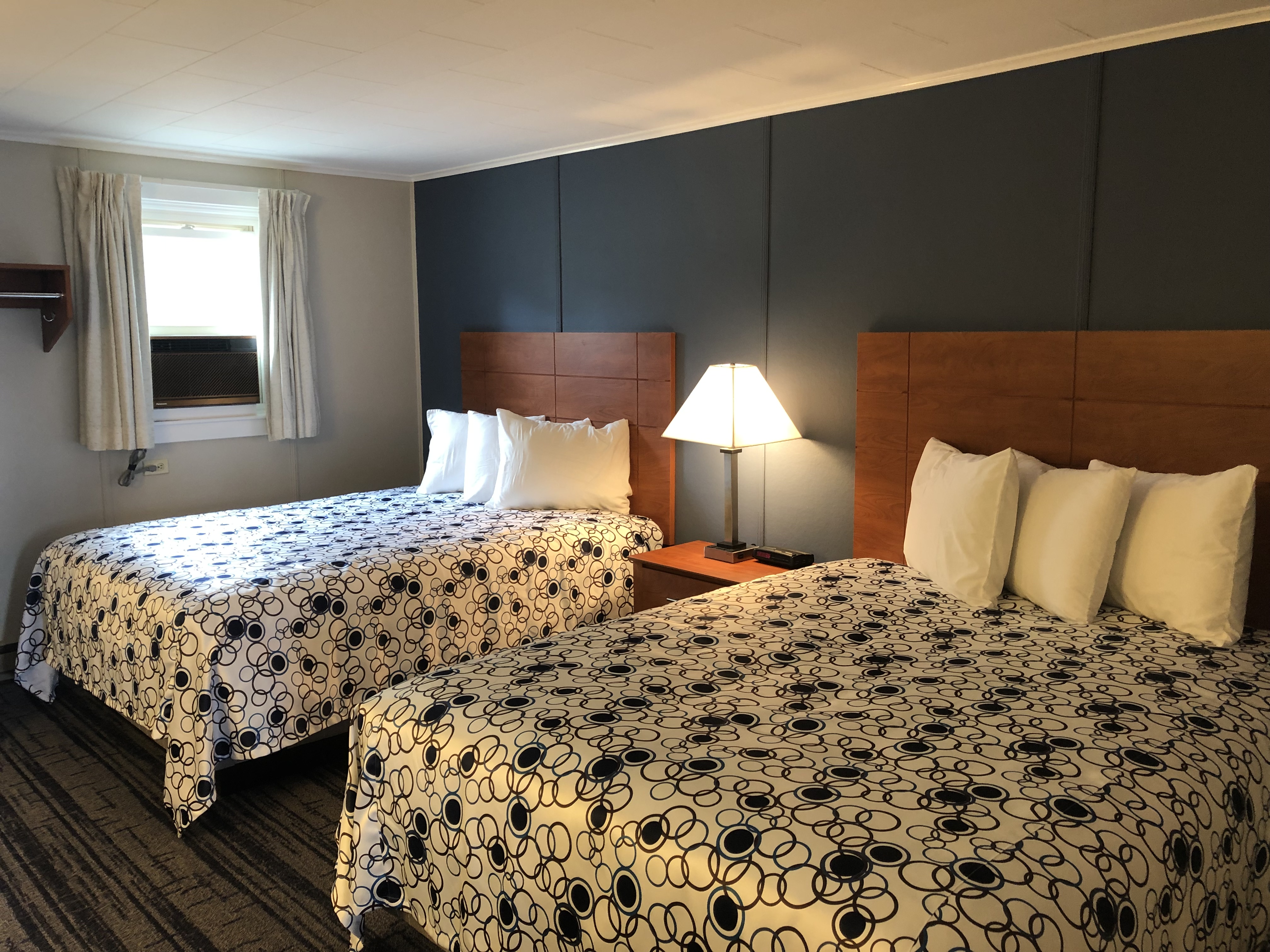 A hotel room with two beds, a nightstand, lamp, and window.